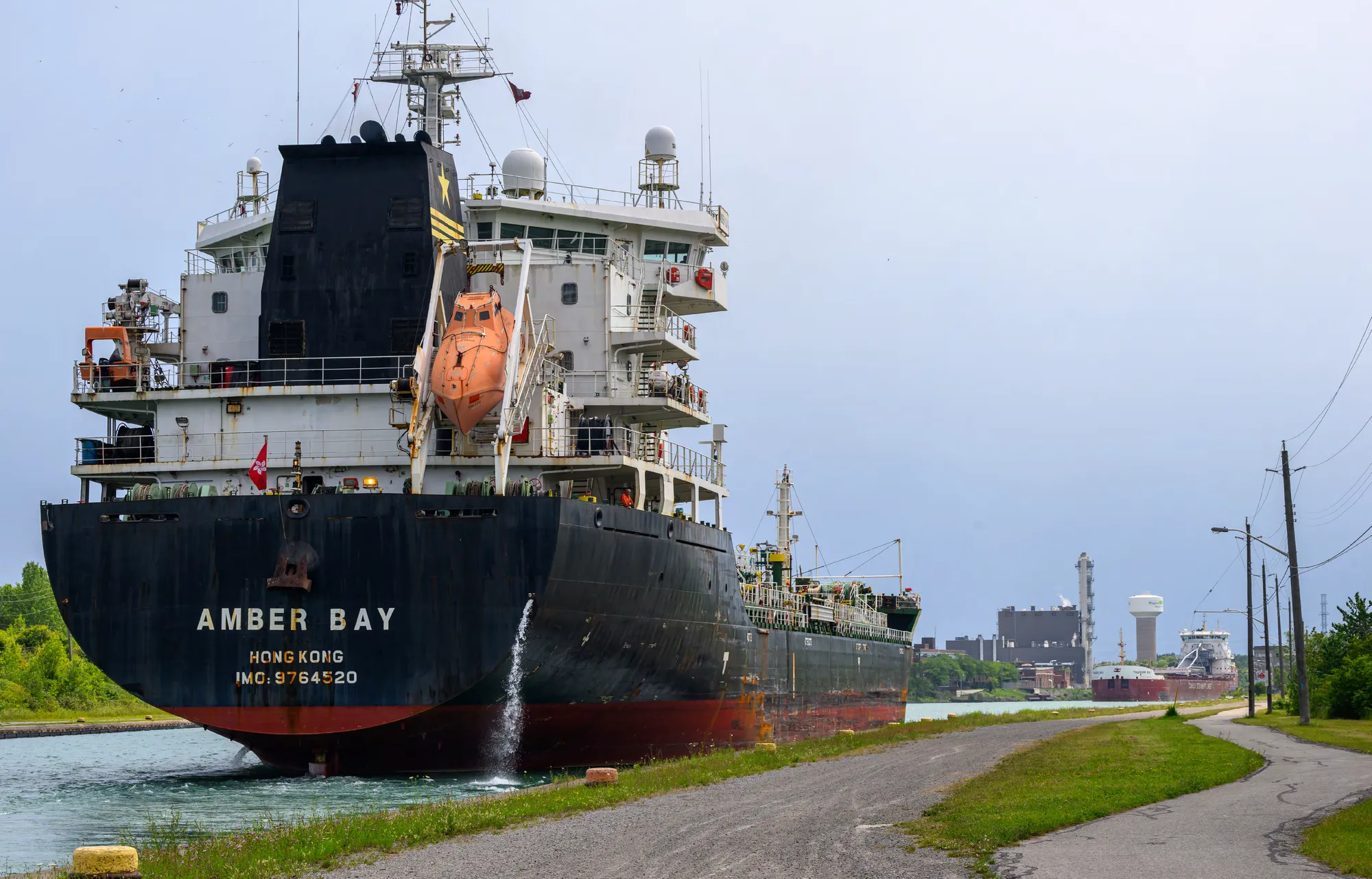Large cargo ship traveling through a shipping canal, with industrial facilities, utility poles, and additional vessels visible in the distance.