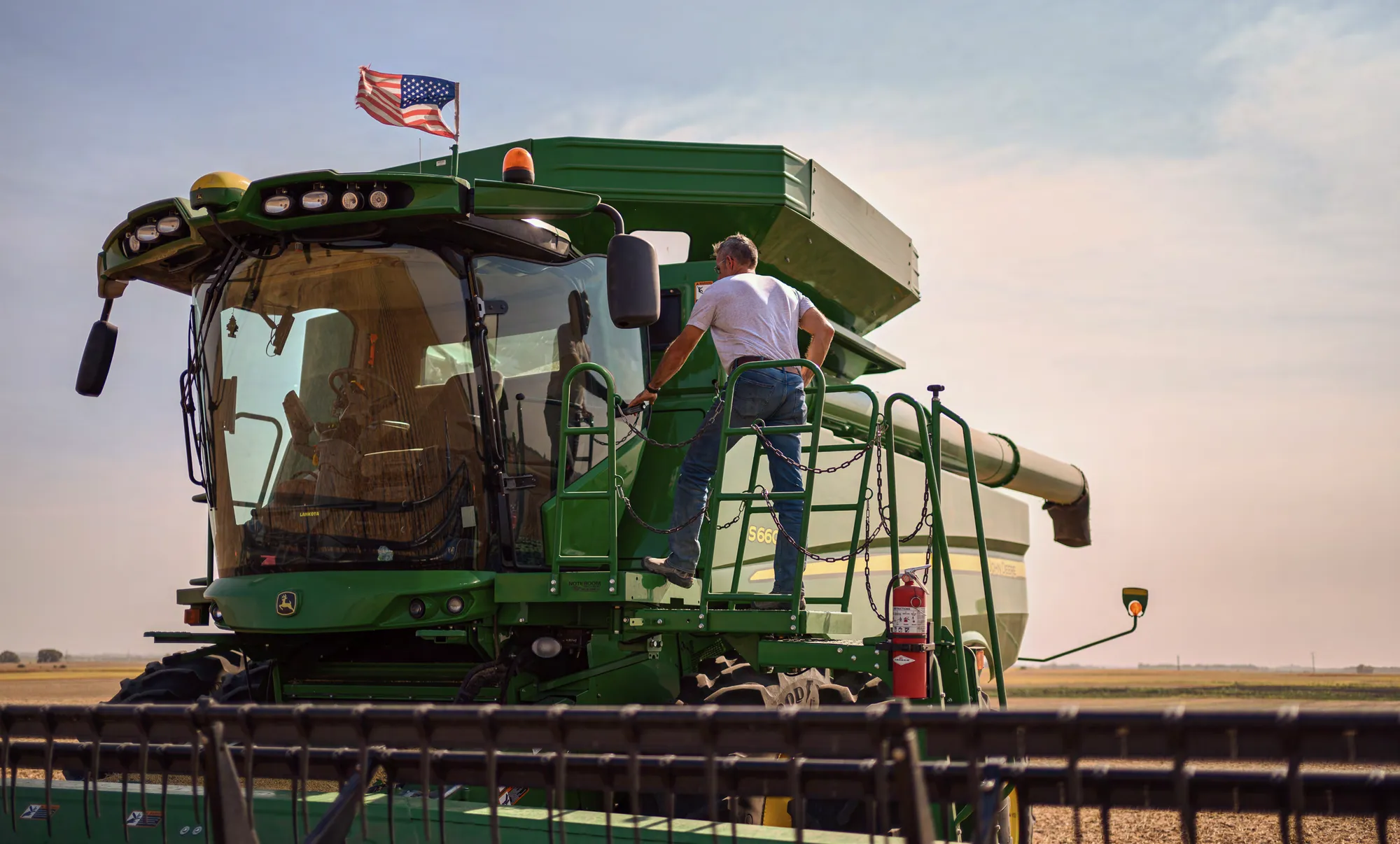 Farmer climbing onto a combine harvester in a field, with an American flag mounted above the cab.