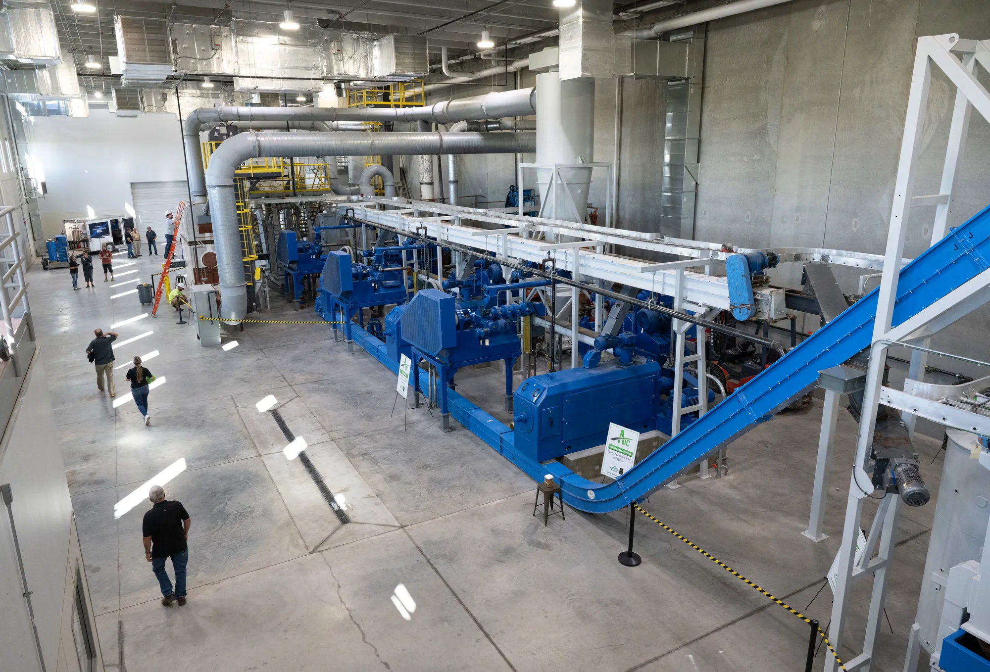 Interior view of a large industrial processing facility with conveyor systems, machinery, overhead ductwork, and workers walking through the production area.