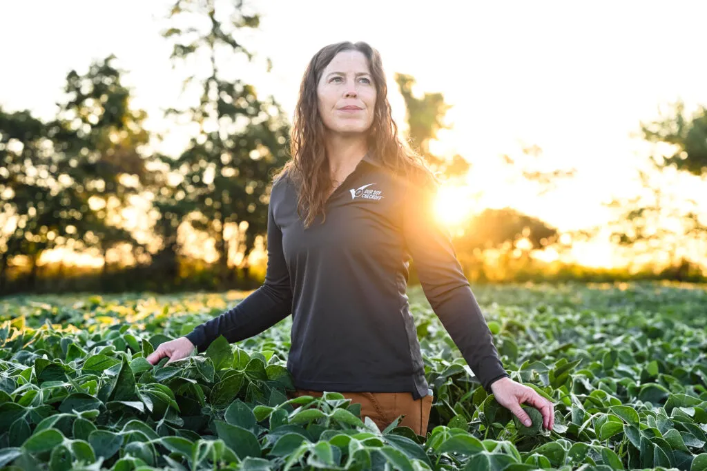 A woman standing in a lush soybean field at sunrise or sunset, with sunlight shining behind her.