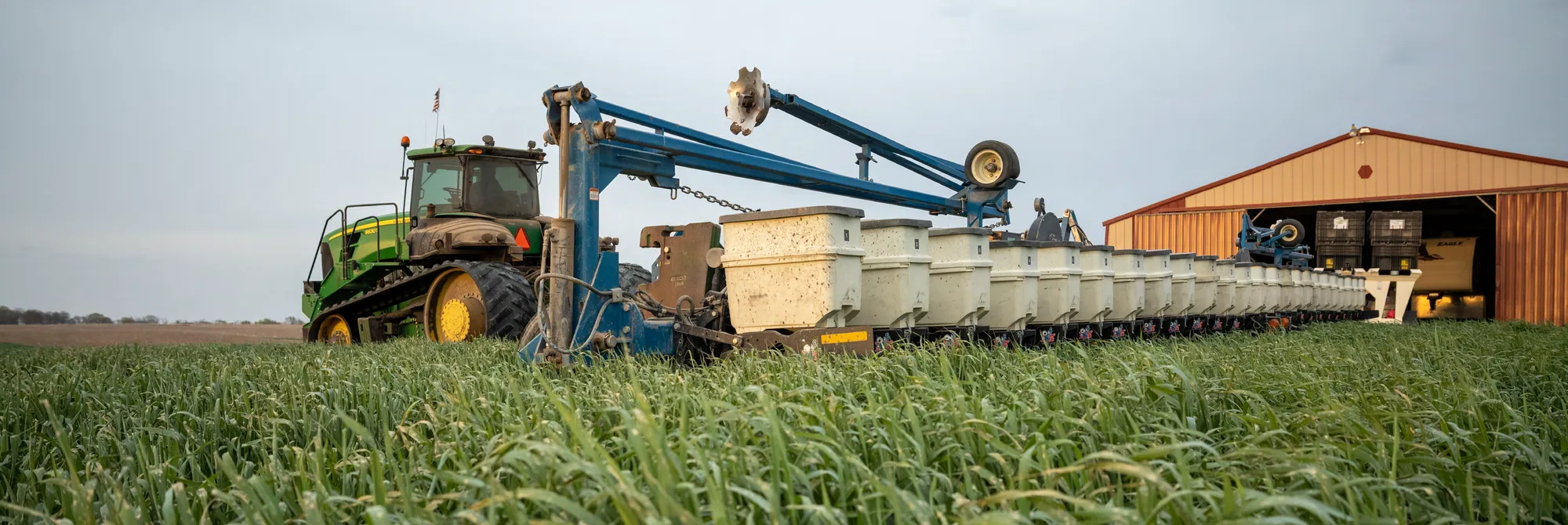 Large agricultural planter positioned in a field of cover crops near a farm building, prepared for planting.