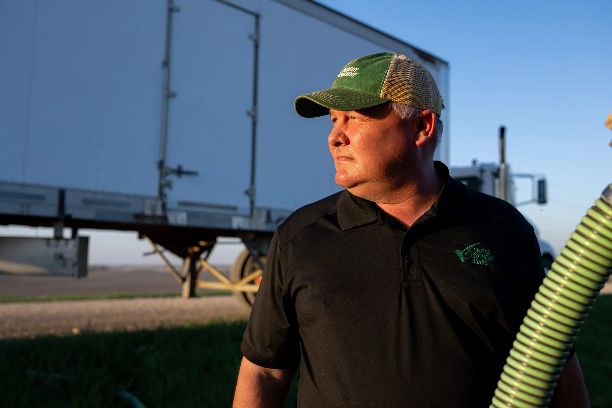 Farmer in United Soybean Board apparel standing near a grain trailer at sunset.