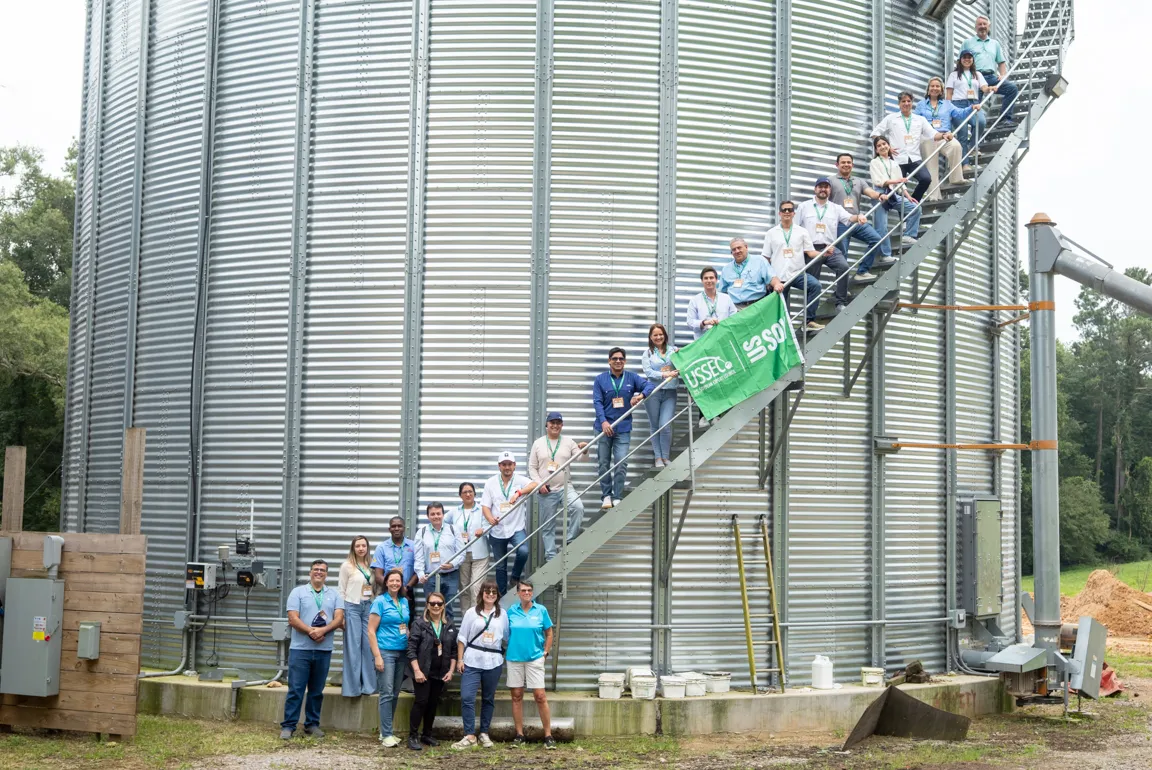 Group of participants pose on and beside a staircase attached to a large grain storage bin, holding a U.S. Soy and USSEC banner.