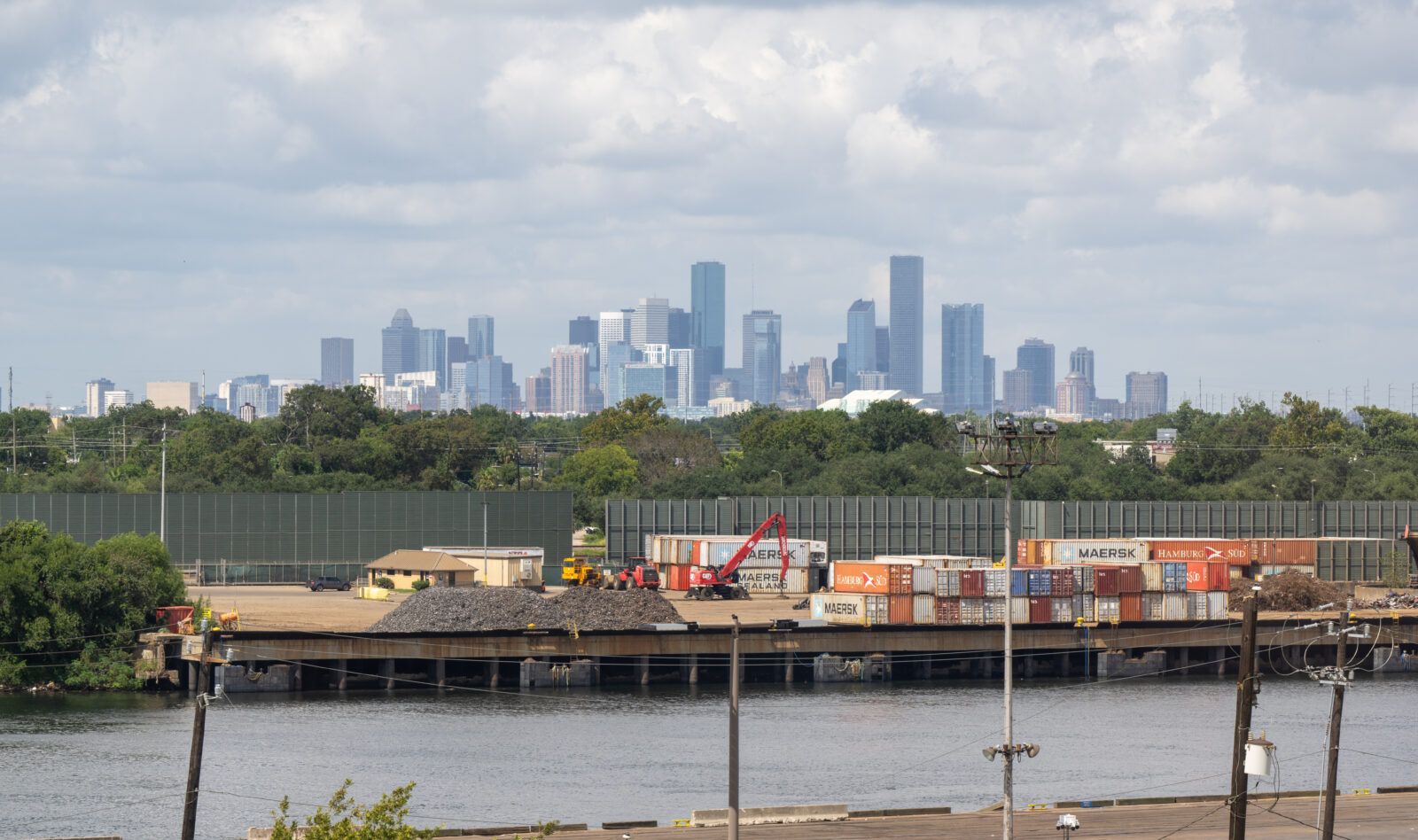 Shipping containers stacked at a port along a waterway, with cranes in the foreground and the Houston skyline in the distance.