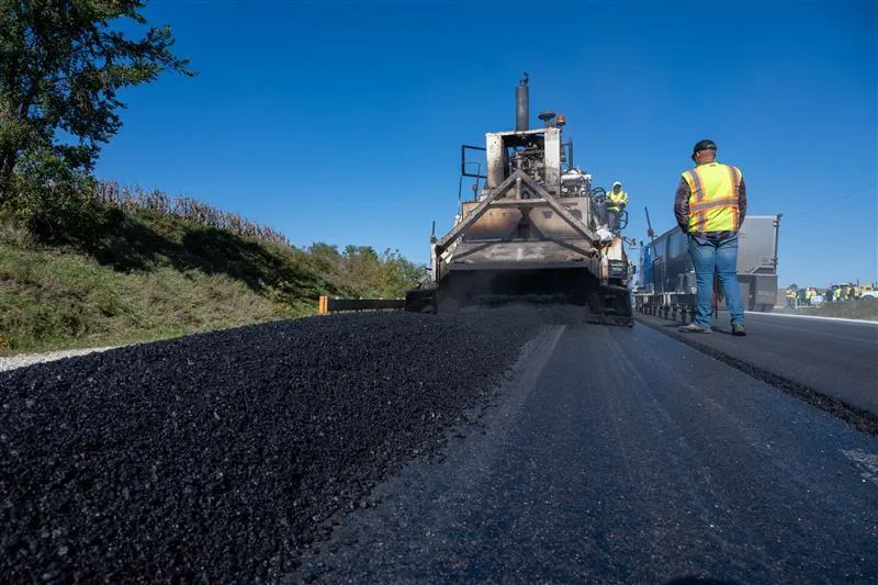 Road construction crew paving a highway, with an asphalt paver laying fresh black pavement while workers in high-visibility safety vests stand nearby under a clear blue sky.