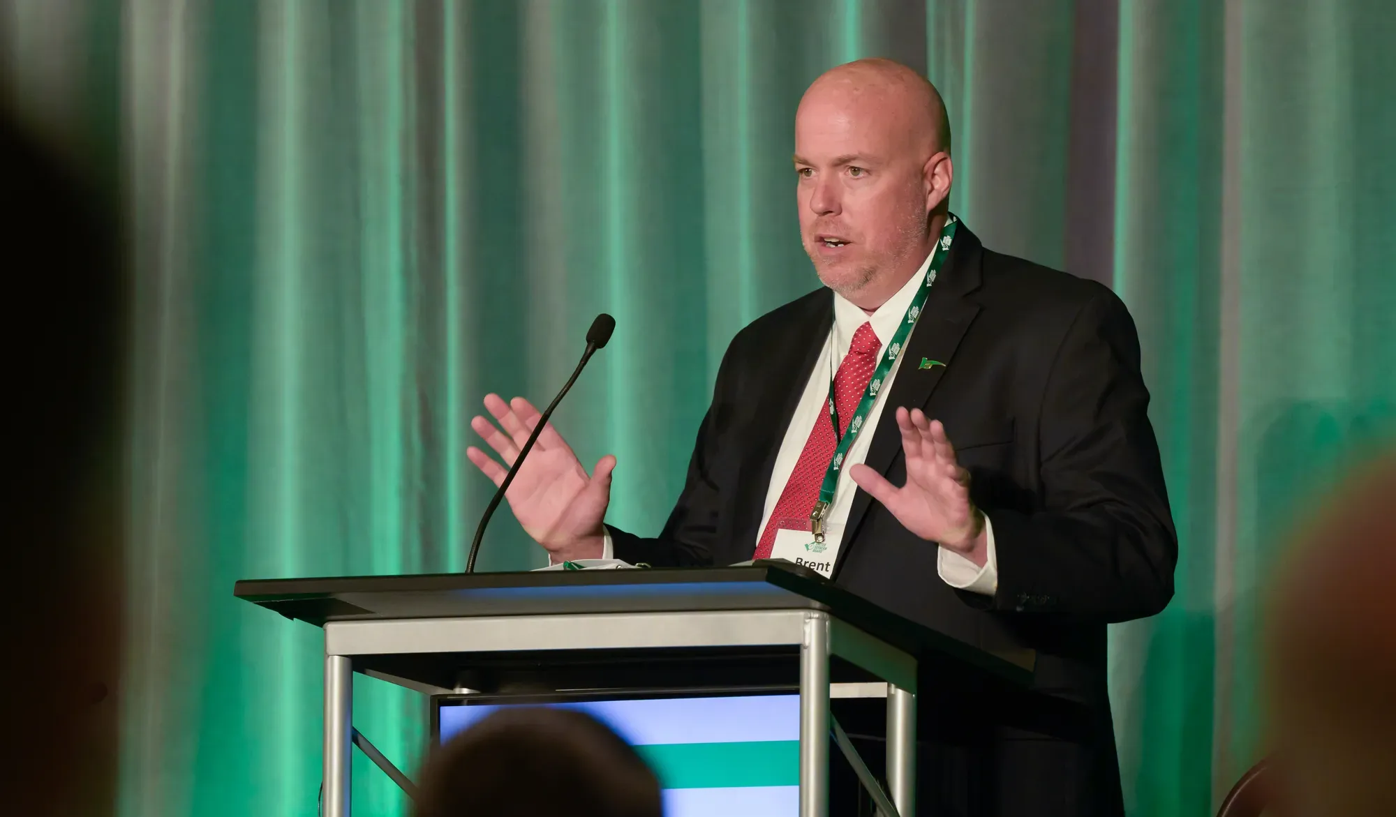 Man speaking at a podium with a green backdrop behind him.