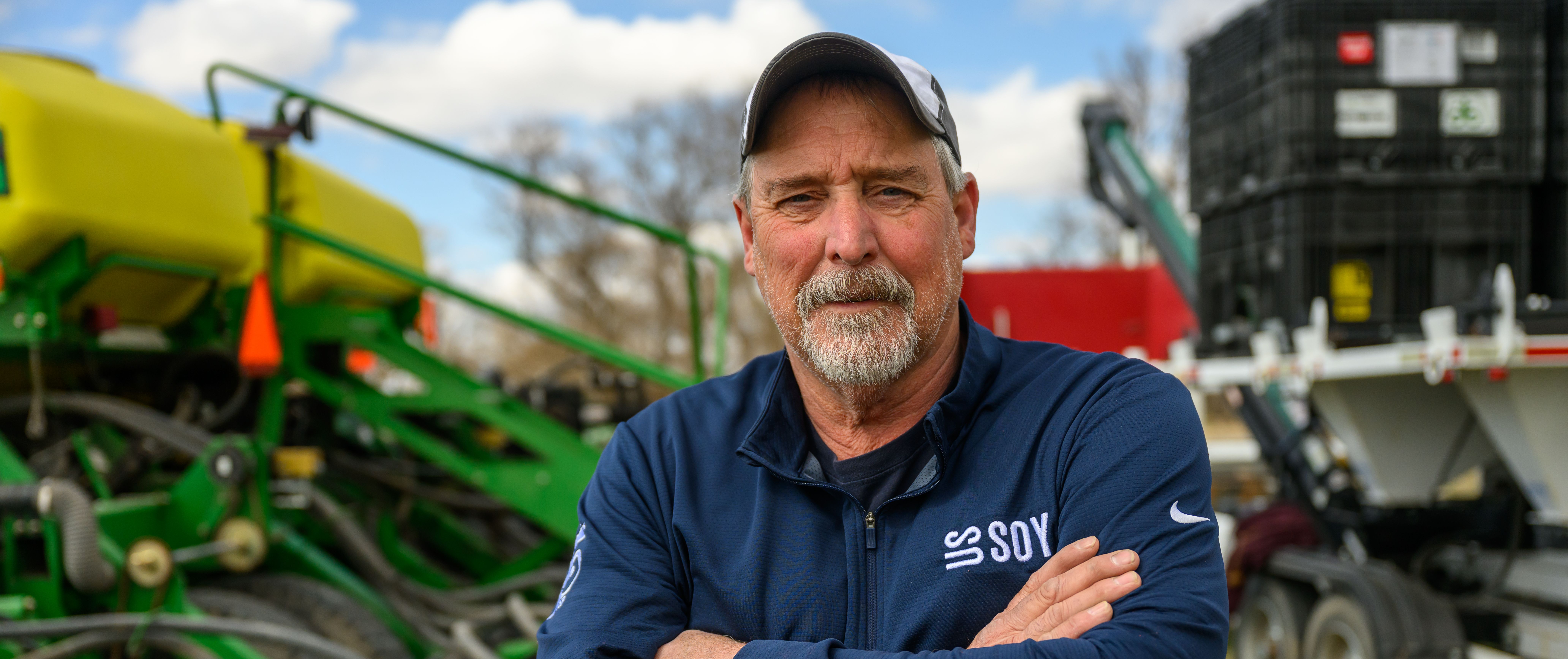 Portrait of a soybean farmer standing with arms crossed in front of planting equipment and machinery in a field.