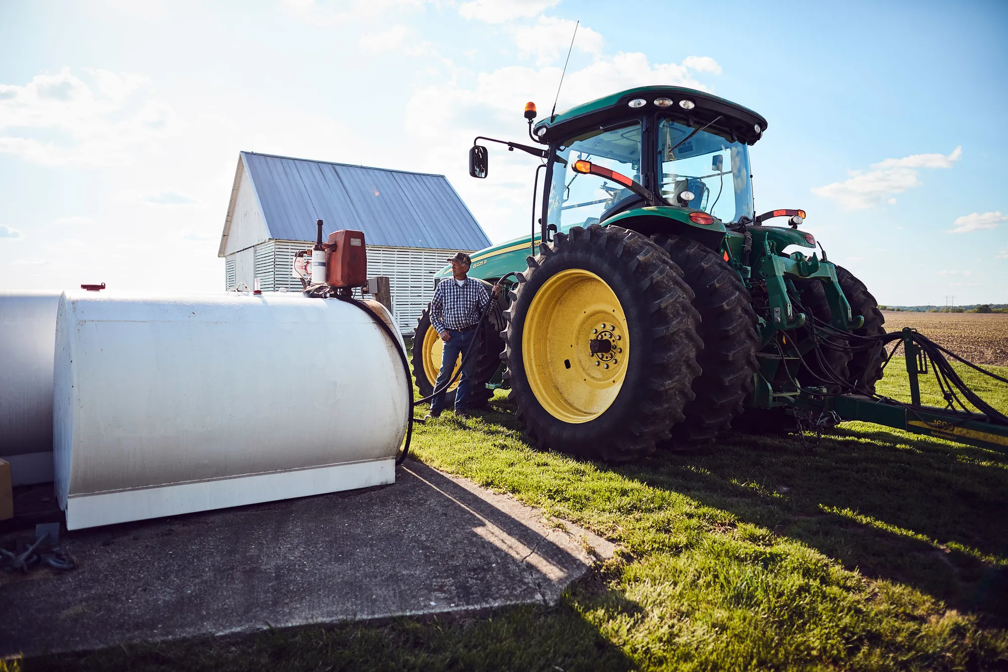 Farmer stands beside a large green tractor near a fuel storage tank and farm building on a sunny day, with agricultural fields in the background.