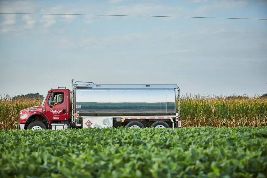 Red tanker truck with a reflective silver tank parked between a green soybean field and a mature cornfield under a blue sky.
