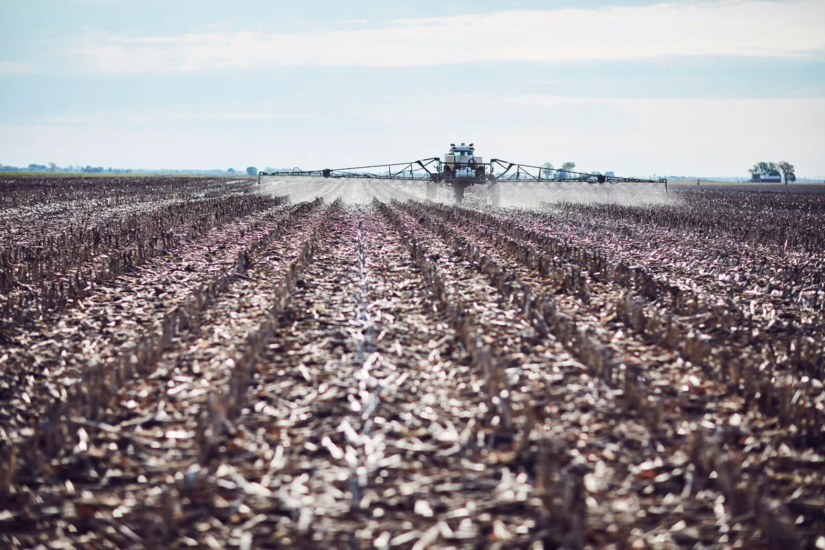 Agricultural sprayer applies a treatment across rows of harvested crop residue in a large field, creating a light mist behind the equipment.