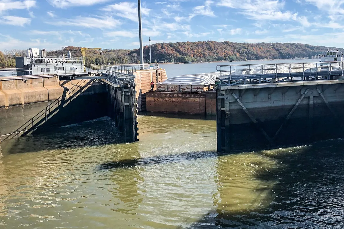 Barge carrying covered cargo passes through a river lock, with wooded hills along the shoreline in the background.