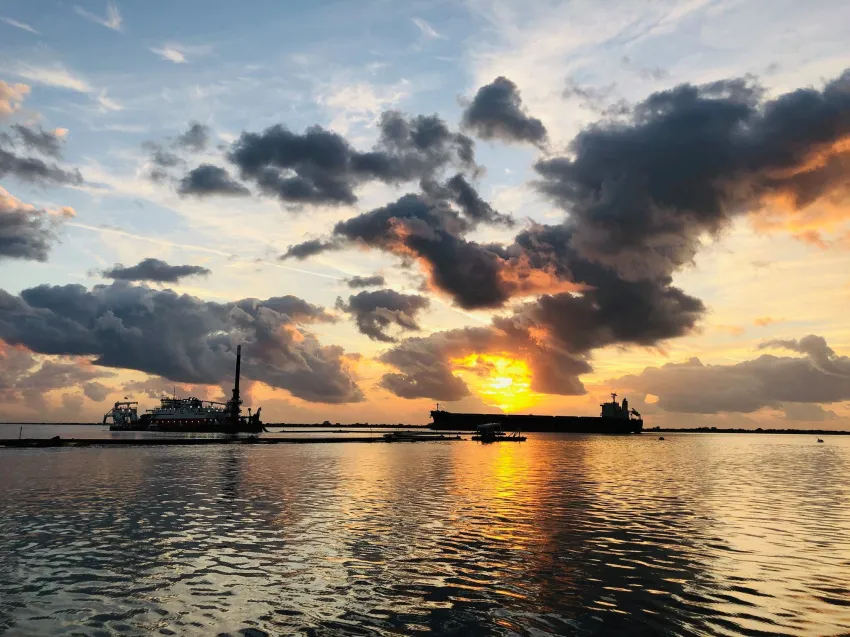 Barges and towboats travel along a river at sunset, silhouetted against dramatic clouds with golden sunlight reflecting across the water.