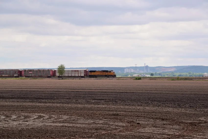 Freight train with a yellow locomotive and railcars travels alongside an agricultural field, with grain storage facilities visible in the distance.
