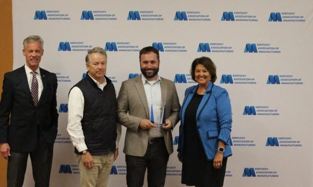 A group of four people standing in front of a Kentucky Association of Manufacturers backdrop, with one person holding an award.