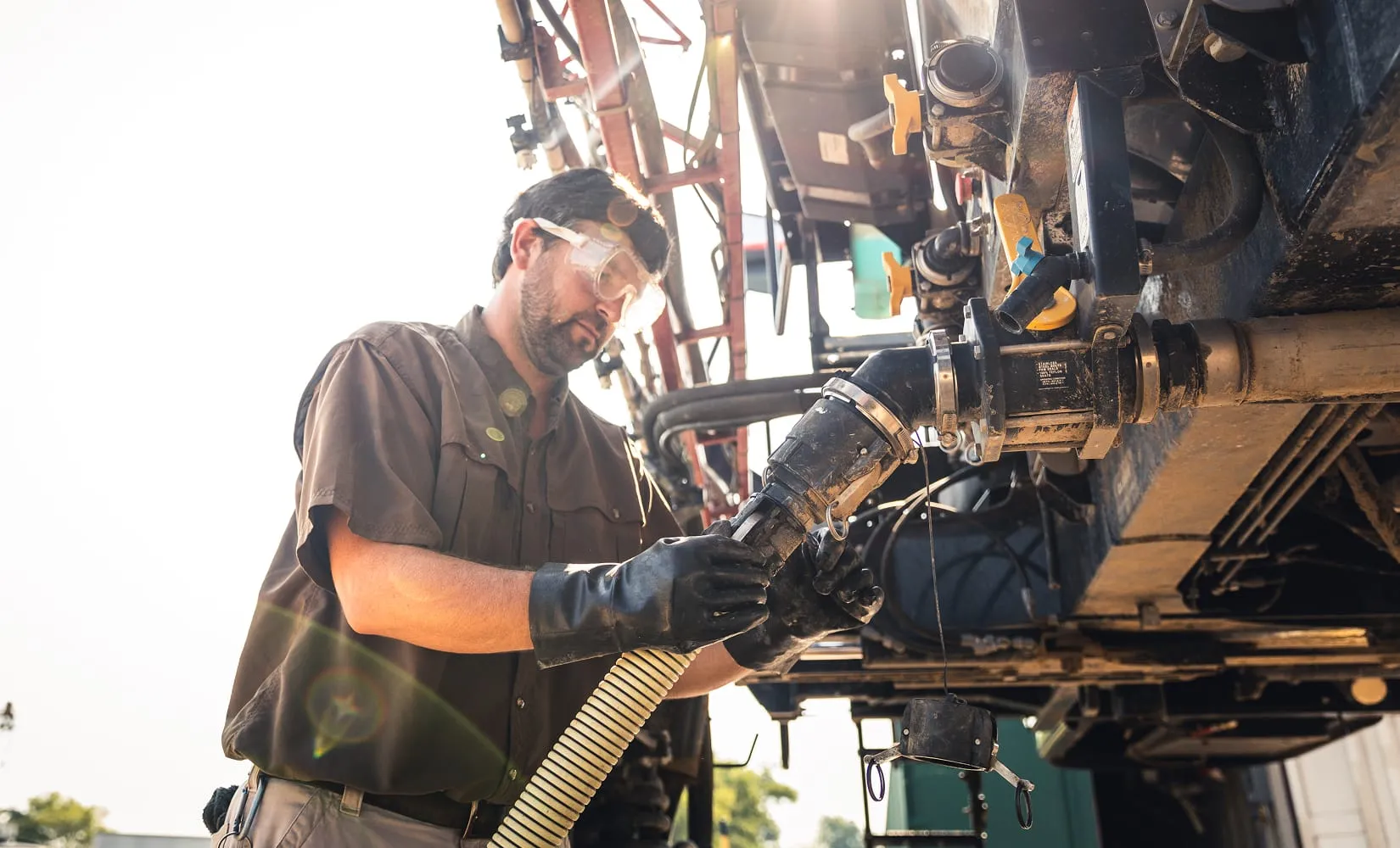 A worker wearing safety goggles and gloves connects a hose to refuel equipment.