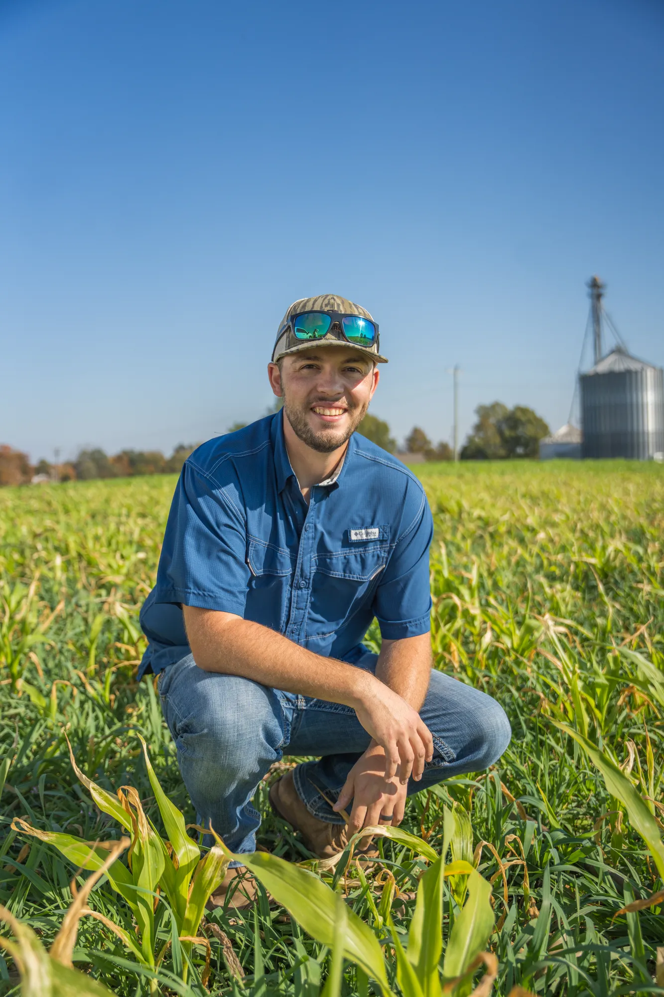 Farmer kneeling in a crop field, smiling toward the camera, with a grain bin and a clear sky in the background.