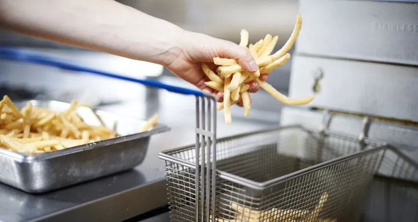 Close-up of a person placing uncooked French fries into a metal fryer basket beside a commercial deep fryer.