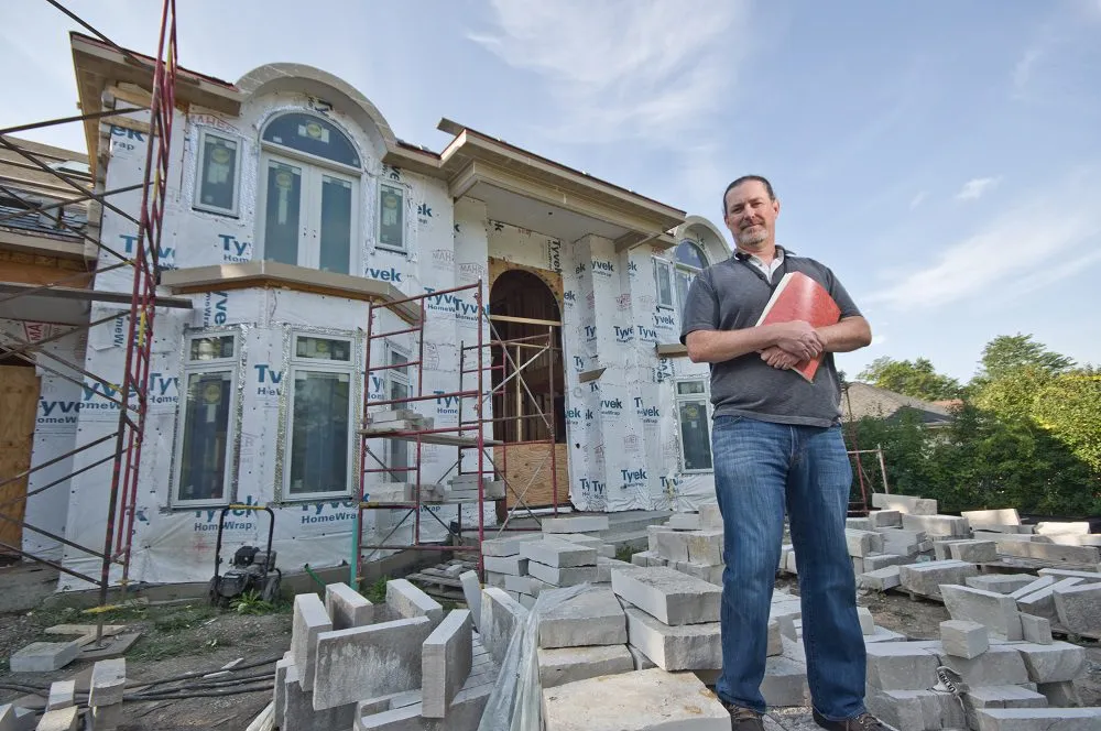 A contractor standing in front of a house under construction, with scaffolding and building materials surrounding the exterior.