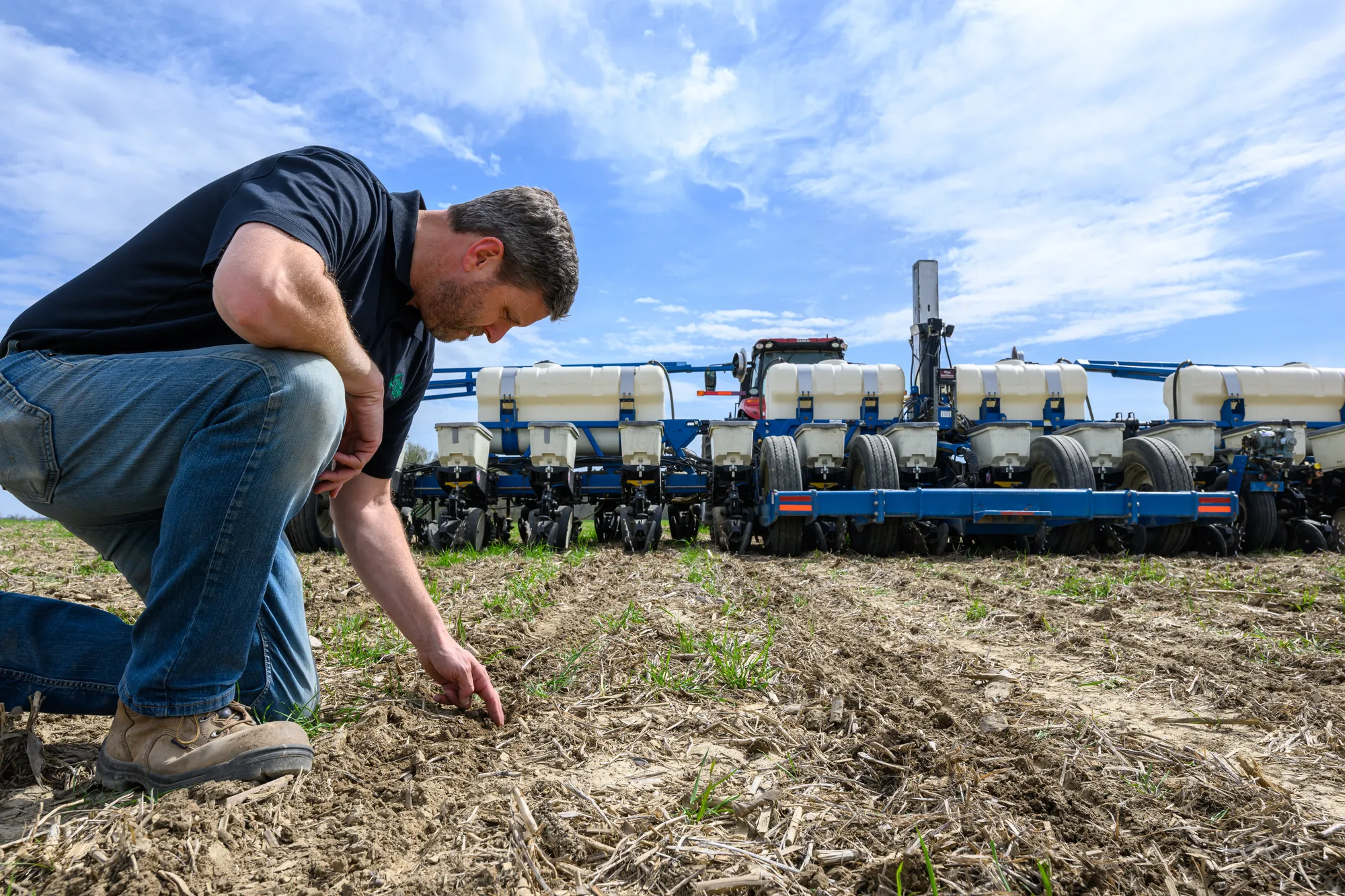 Farmer kneeling in a field to inspect emerging soybean crops, with a large planter in the background.