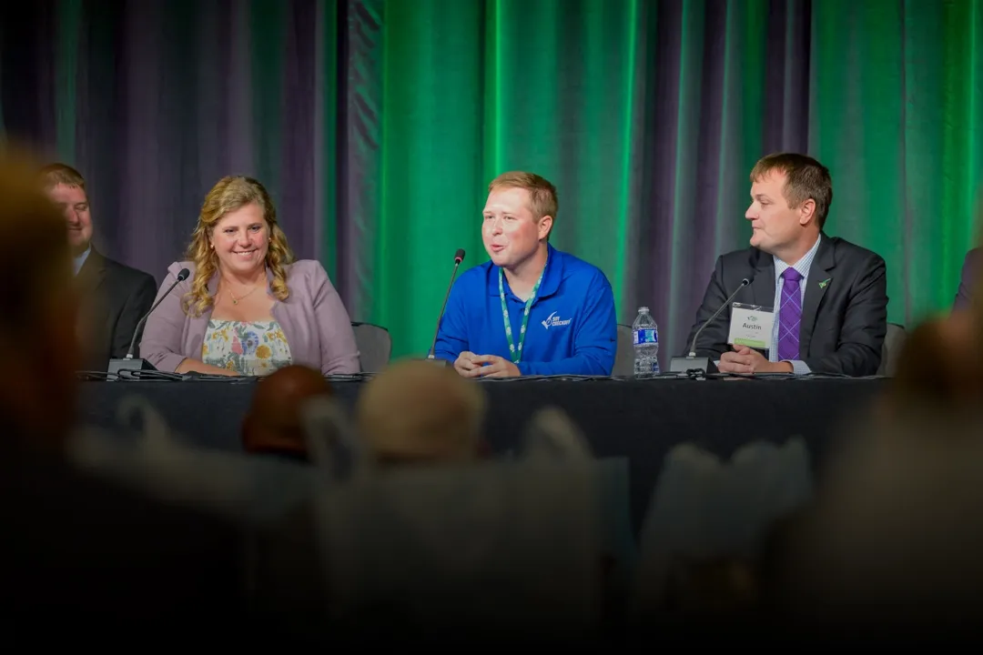 Three panelists seated behind microphones speak at a conference table, with audience members in the foreground and green-lit curtains behind them.
