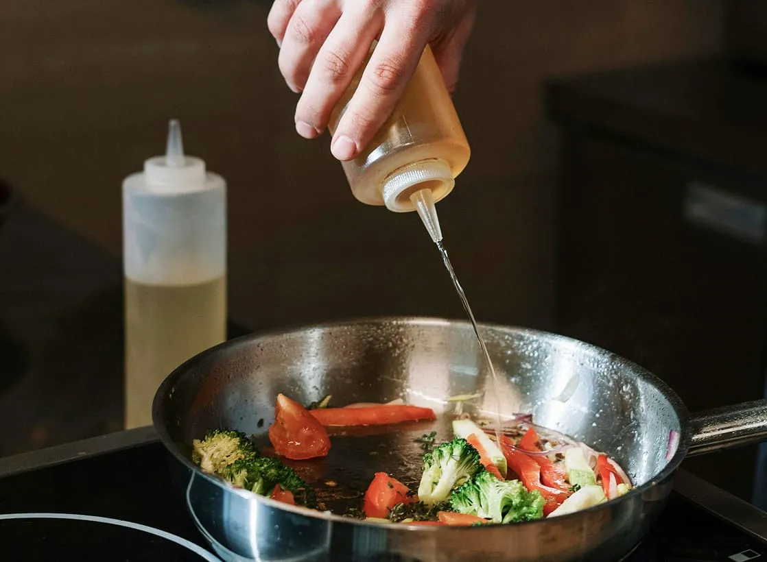 A hand drizzles oil from a squeeze bottle into a pan of sautéing vegetables.