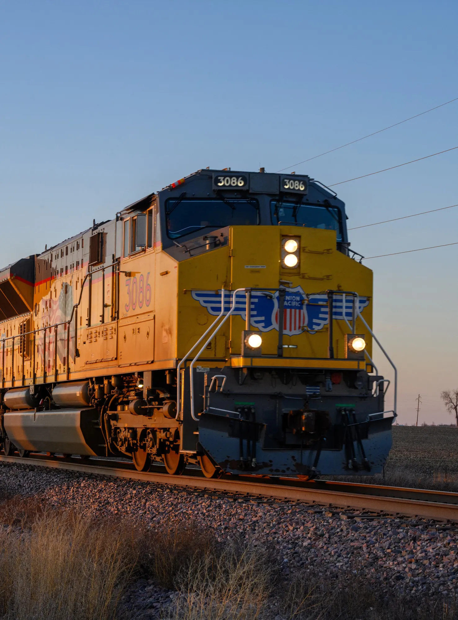 A powerful yellow freight train pulls tank cars along a rural track at sunset.