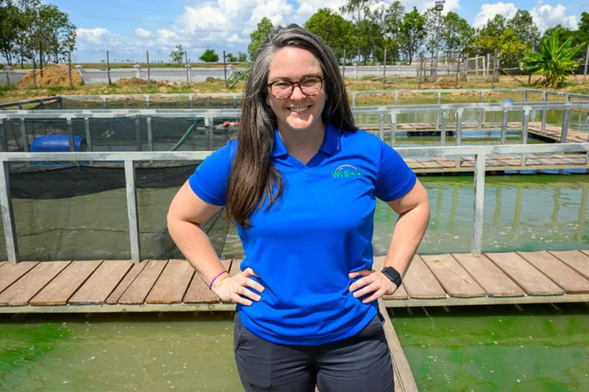 A woman in a WISHH shirt stands beside aquaculture ponds at a fish farming facility.