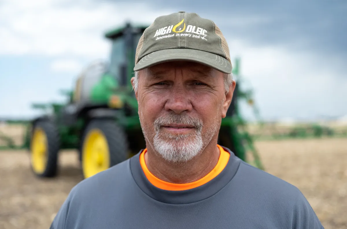 Soybean farmer wearing a High Oleic cap stands in a field with a tractor in the background.