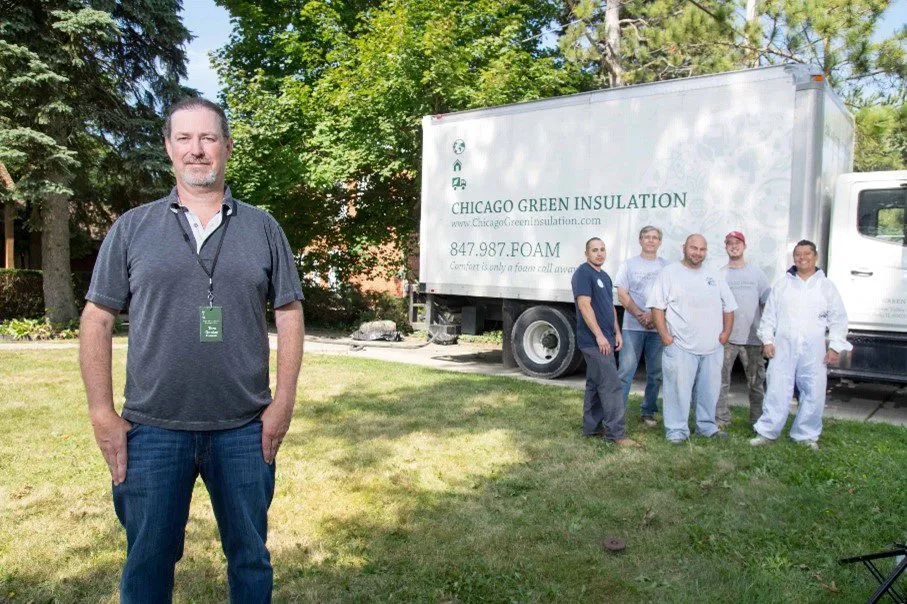 A man stands in the foreground while a crew poses beside a truck labeled ‘Chicago Green Insulation’ parked on a lawn.