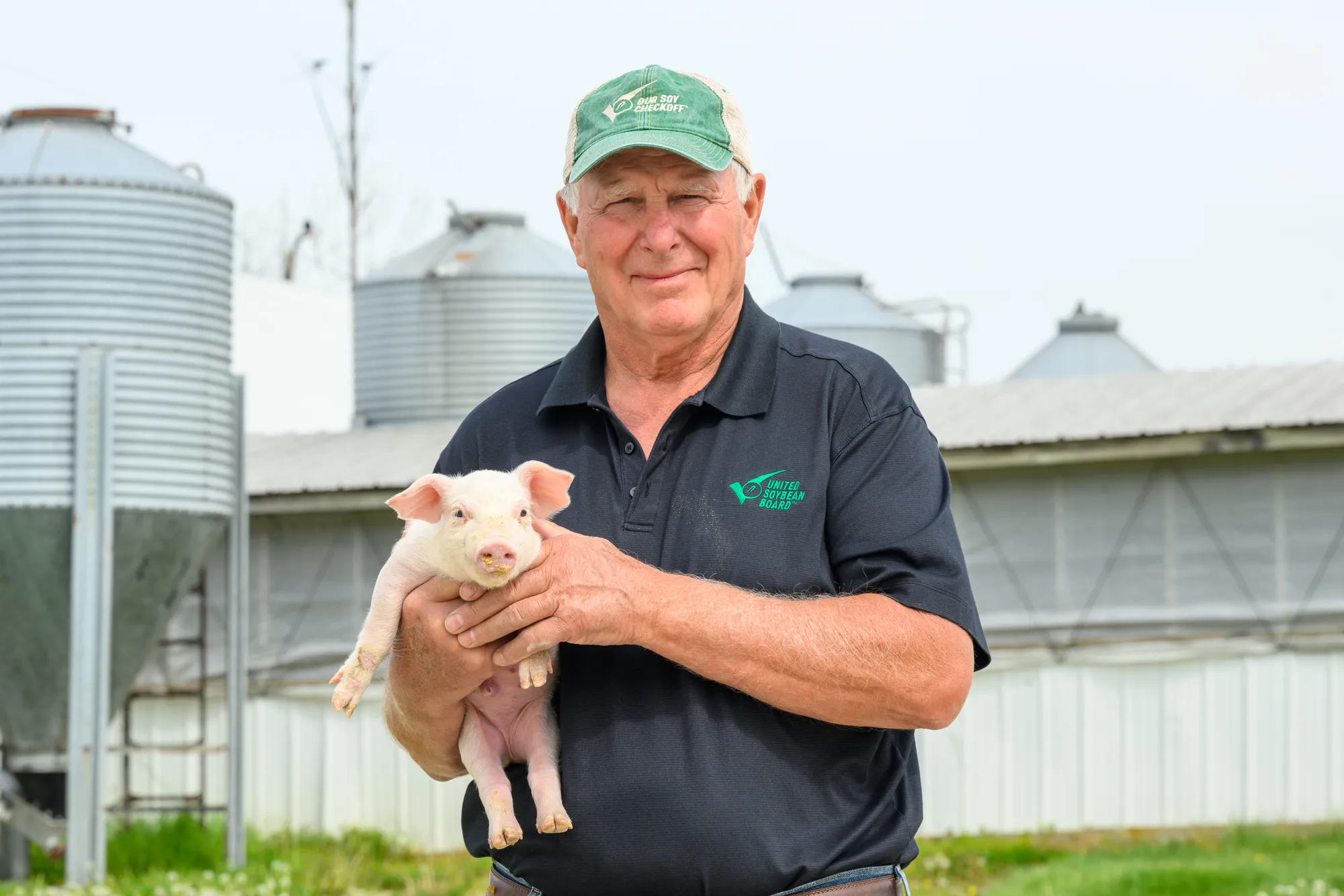 Farmer holding a piglet outdoors near farm buildings and grain bins.