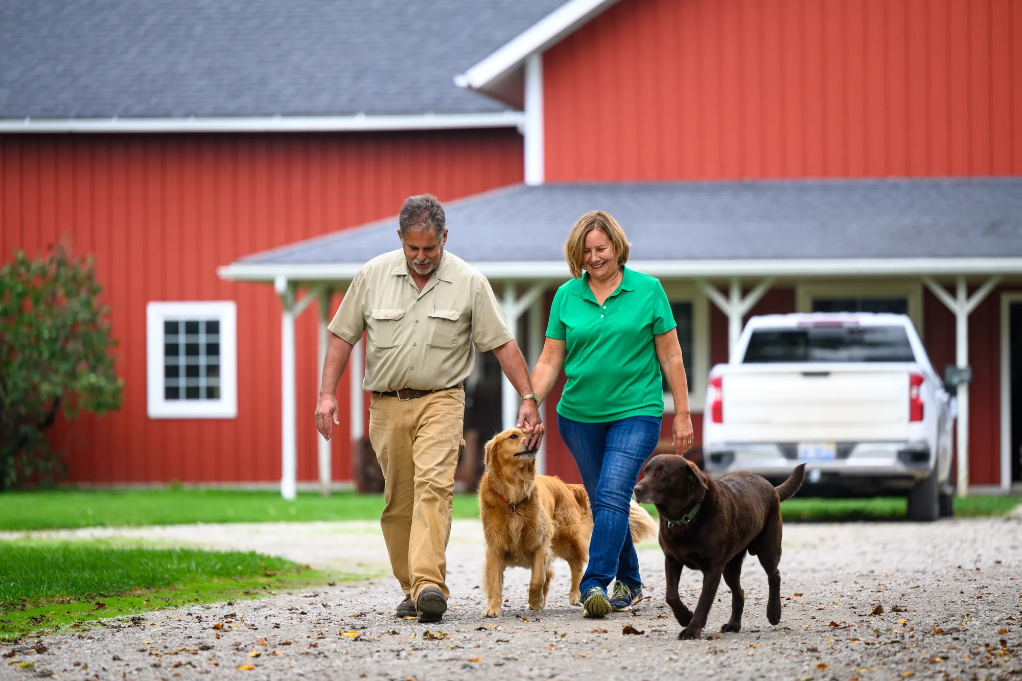 Husband and wife walking two dogs along a gravel driveway in front of a red building, creating a rural farmstead scene.