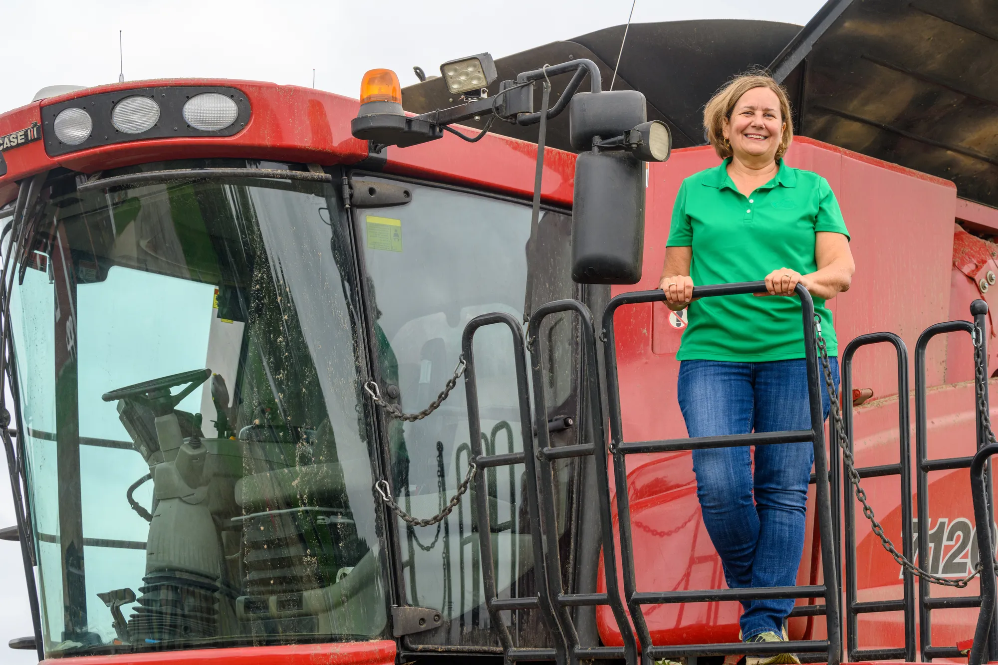 Michigan farmer standing on the platform of a combine harvester, smiling while holding the railing.