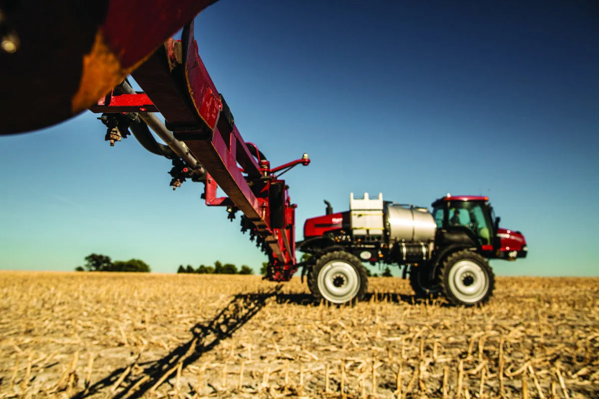 Agricultural sprayer with an extended boom in a harvested field under a clear blue sky.