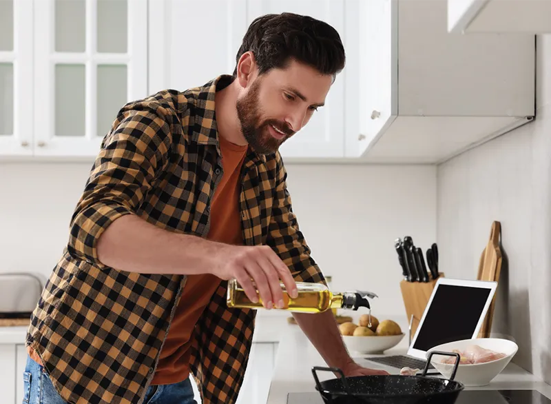 A man in a plaid shirt pours cooking oil into a pan on the stove while preparing a meal in a kitchen