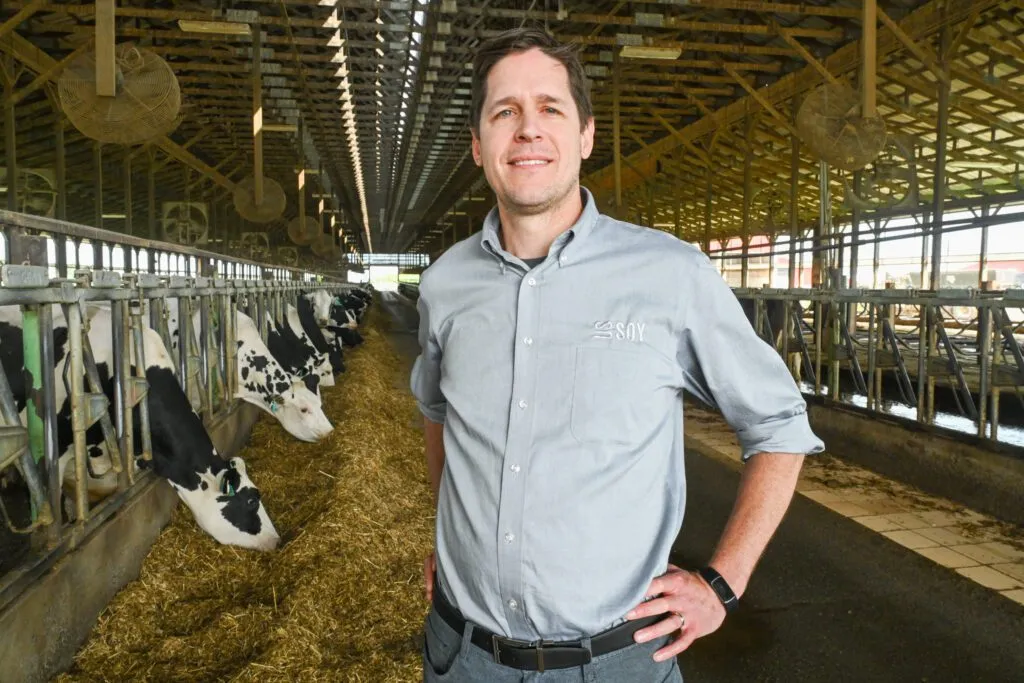 A man stands in a dairy barn, smiling as cows feed behind him.