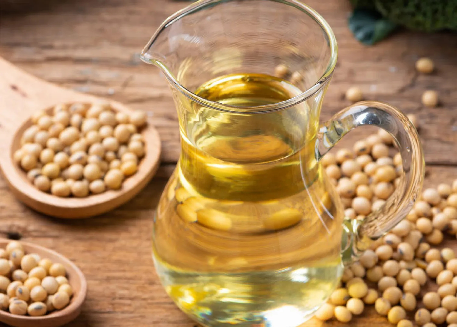 Glass pitcher of soybean oil on a wooden table surrounded by soybeans and green vegetables.