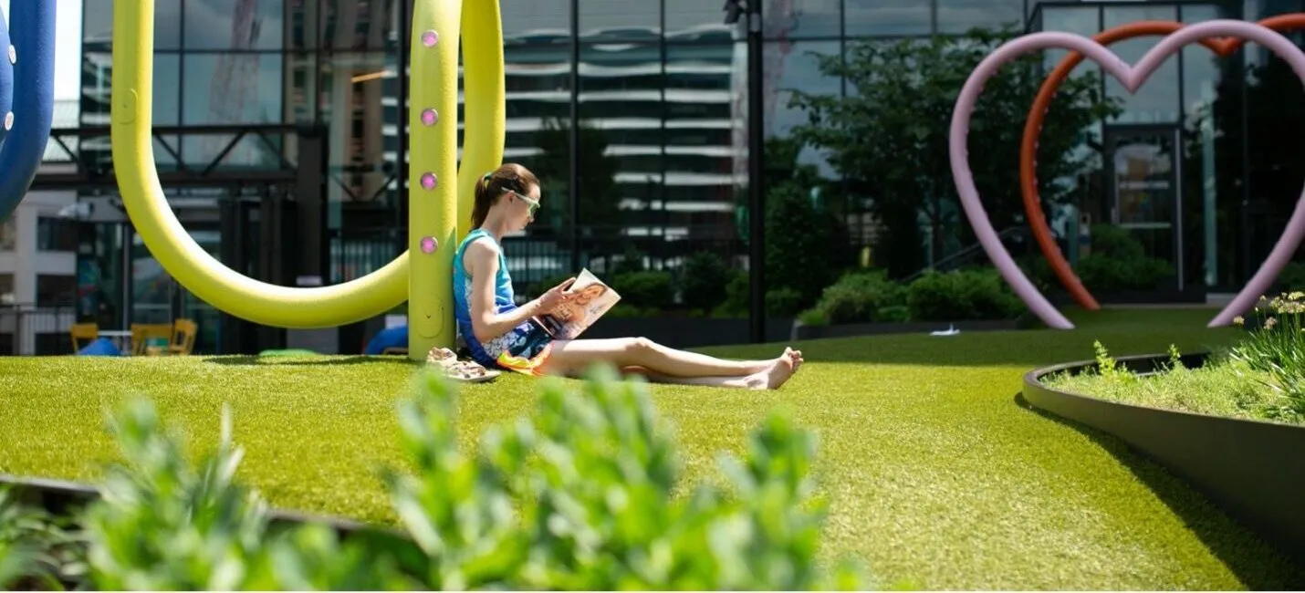 A person sitting on artificial grass, leaning against a colorful playground structure while reading a magazine in an outdoor urban space.
