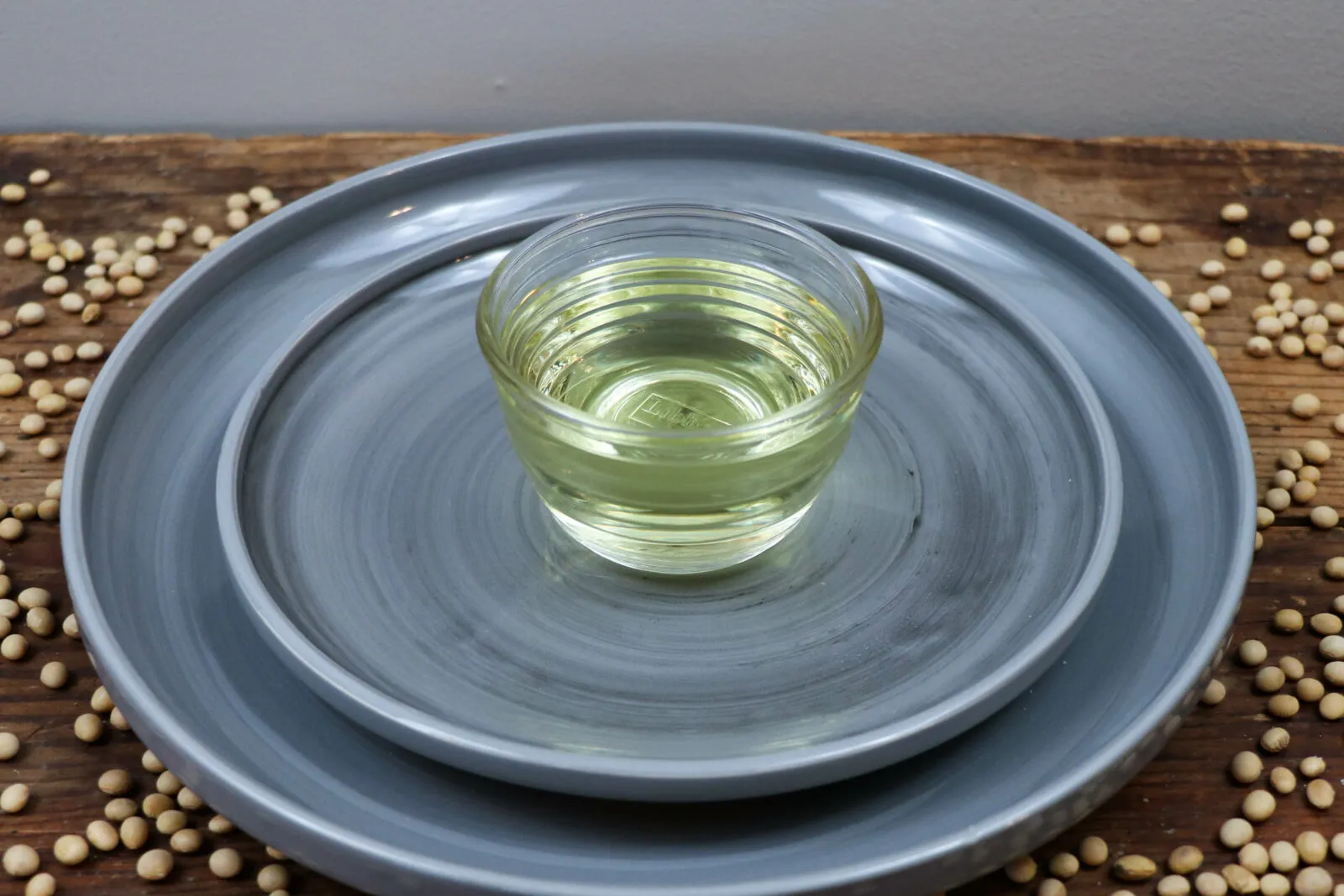 Small glass bowl of soybean oil on stacked gray plates, surrounded by whole soybeans on a wooden surface.