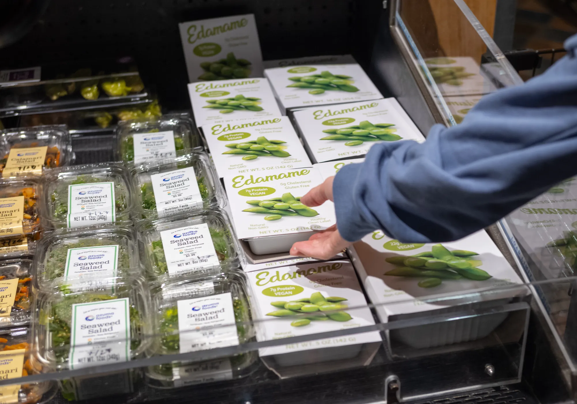 Shopper reaching for a package of edamame displayed in a supermarket refrigerated case.