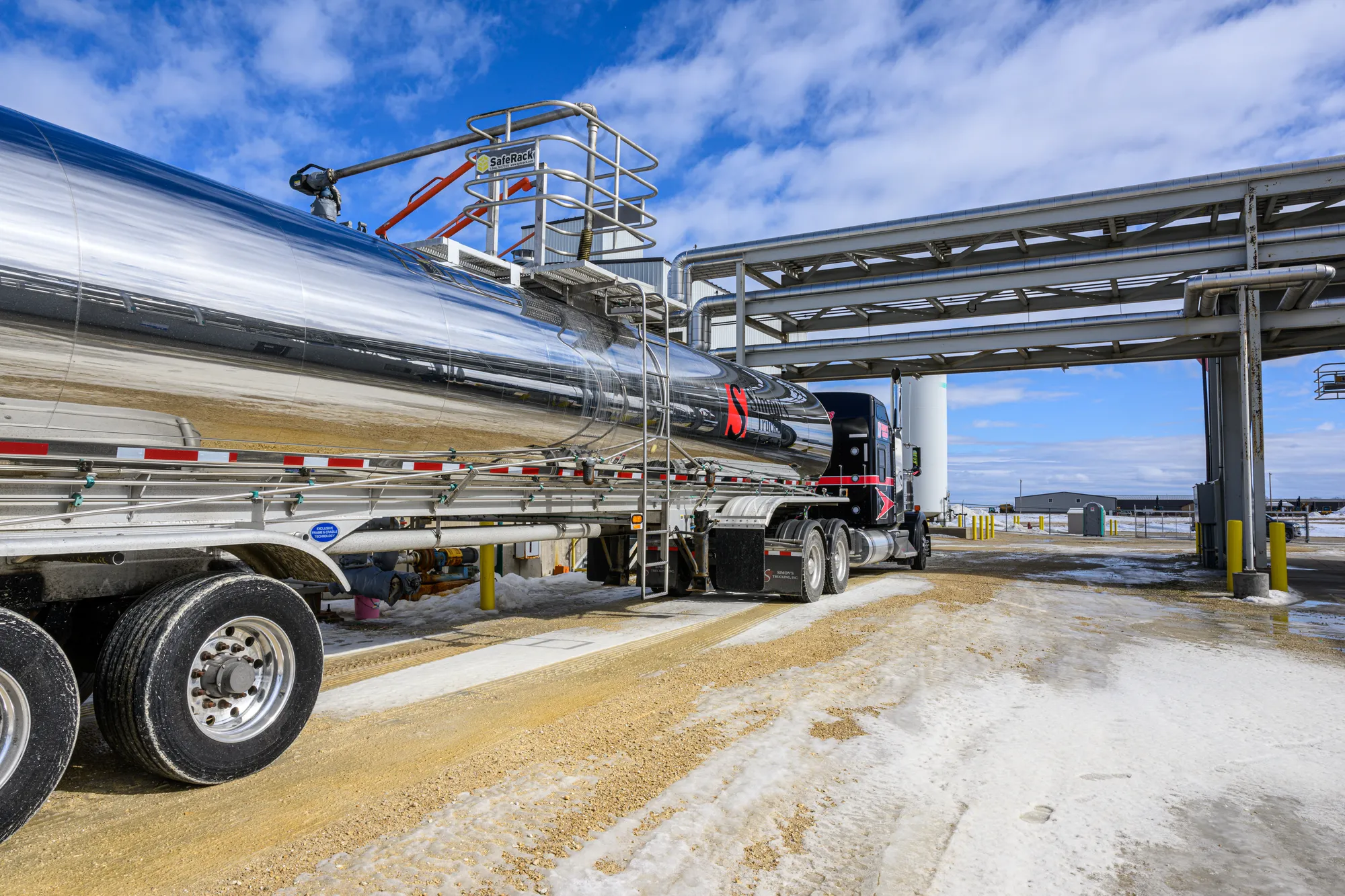 Truck positioned at an industrial loading facility, with overhead piping and transfer equipment connected for fuel or liquid transport.