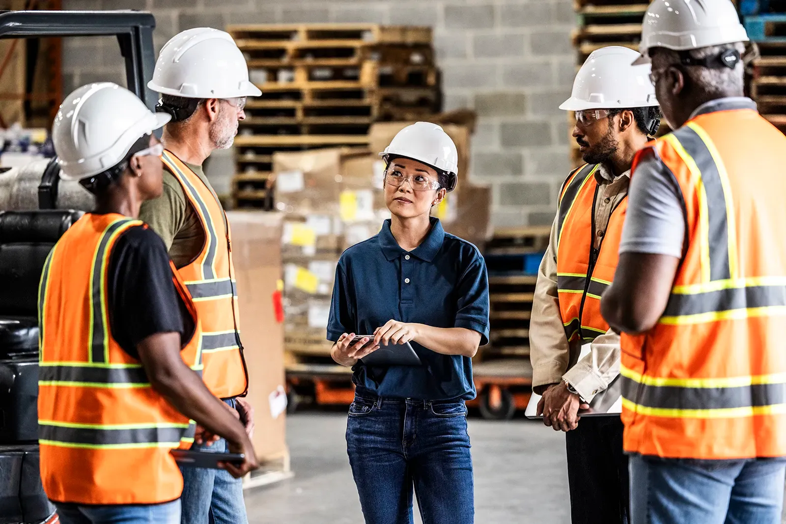 A group of warehouse workers wearing safety helmets and reflective vests listen attentively to a consultant holding a tablet, who appears to be providing guidance in an industrial setting.