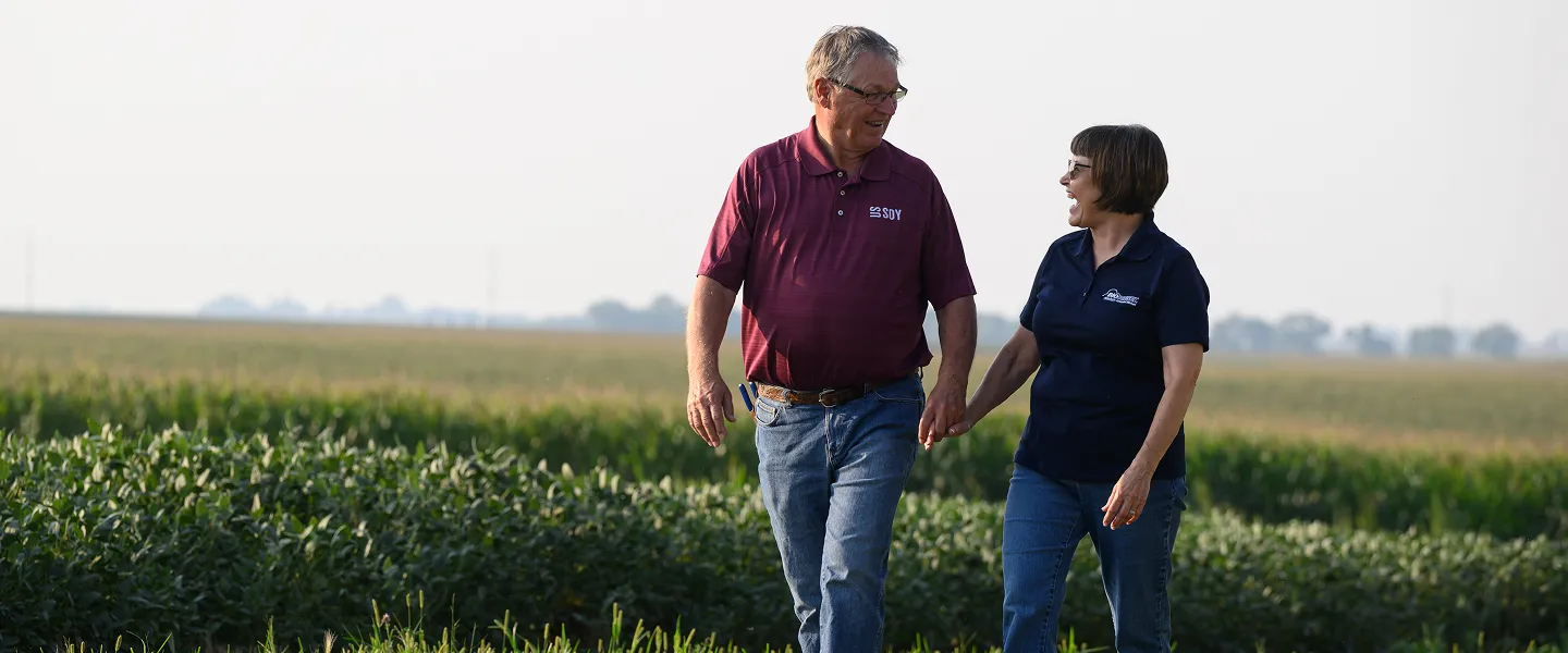 Husband and wife walking together along the edge of a soybean field, talking as they pass rows of growing plants.