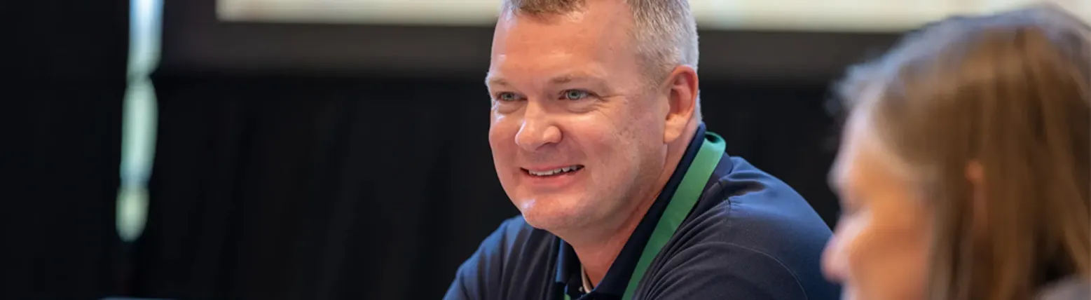 Man in navy polo and green lanyard smiling during a conversation at a table with a laptop.