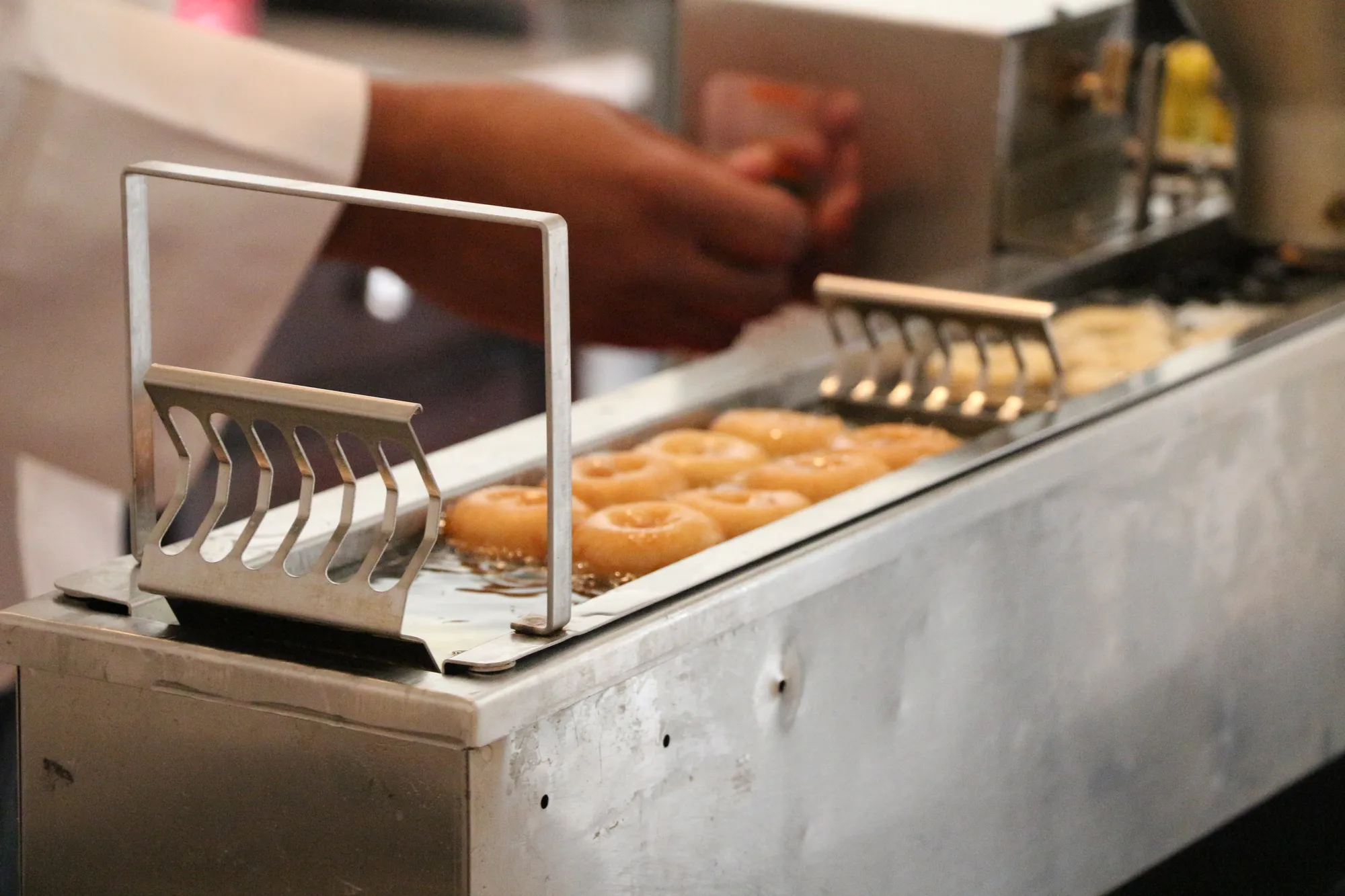 Rows of donuts frying in high oleic oil as a worker tends the commercial fryer.