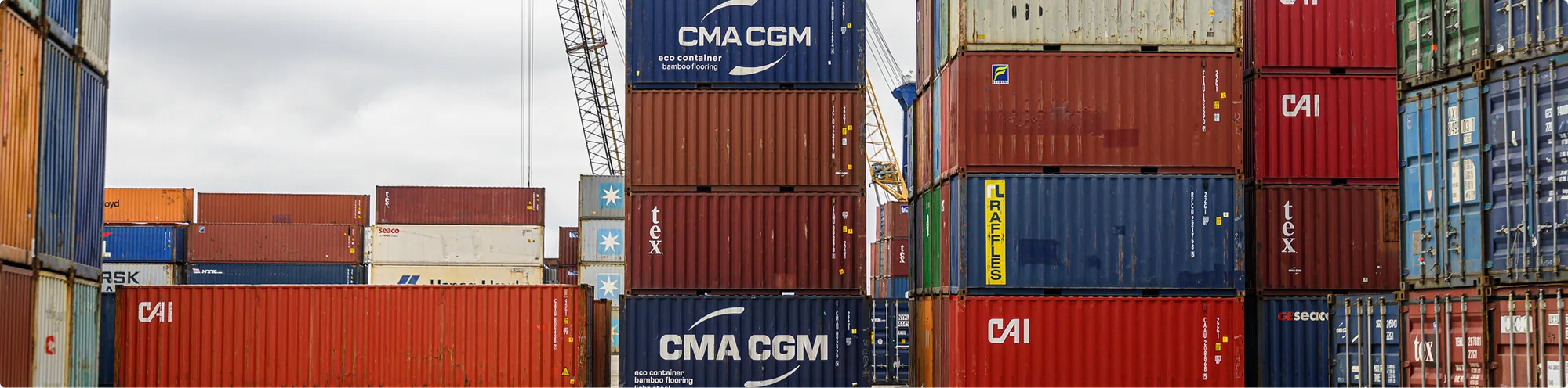 Stacks of shipping containers in a cargo yard at a port or logistics facility, with cranes and industrial infrastructure visible in the background.
