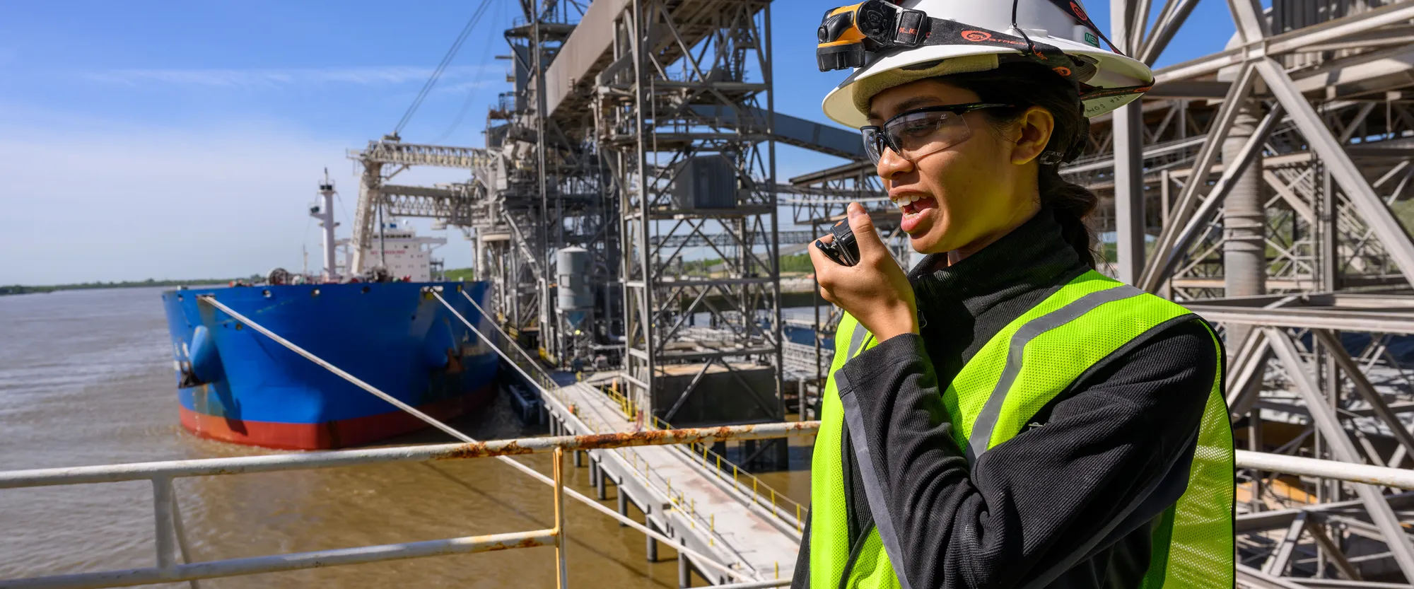 Worker wearing a hard hat, safety glasses, and a vest using a radio at an industrial port facility, with a cargo ship docked alongside the loading infrastructure.