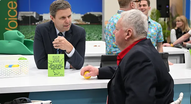 Two men talking at a table at a conference or event, with other attendees visible in the background.