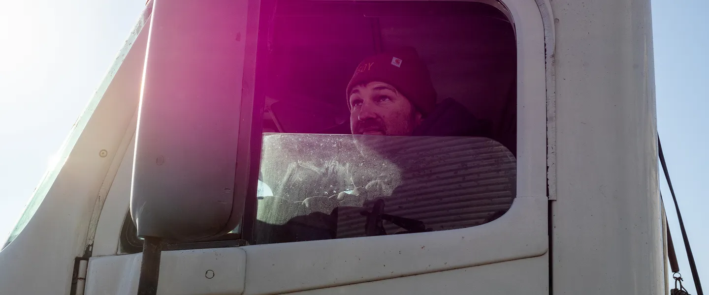 Farmer seated inside a truck cab, looking out the side window, with sunlight streaming through the windshield.