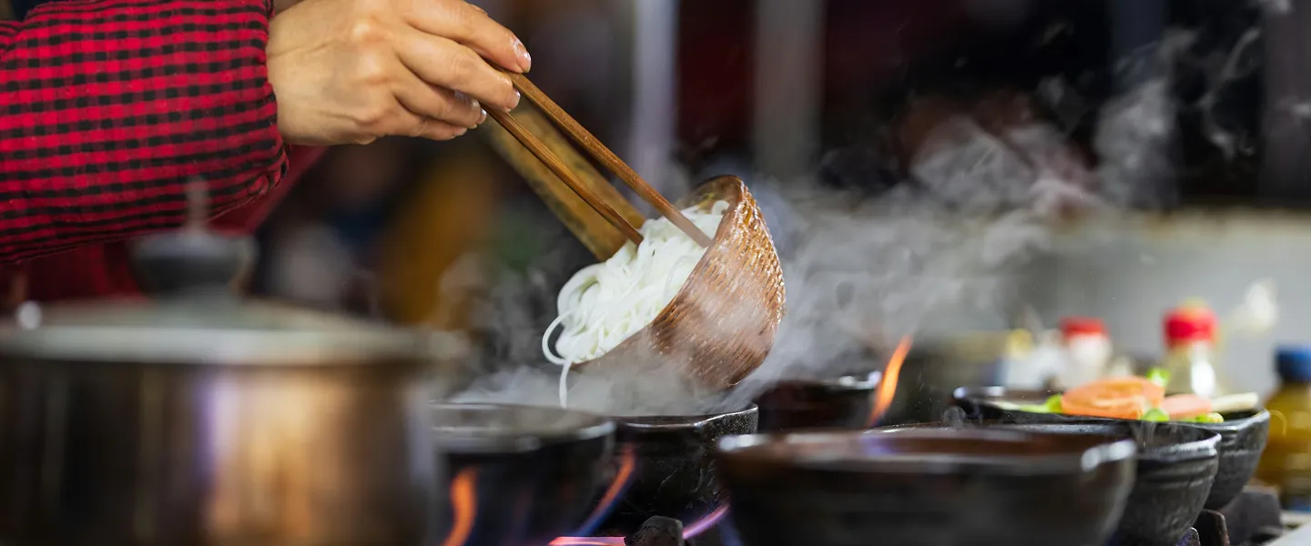 A close-up of a person using chopsticks to lift noodles into a pan with soybean oil.