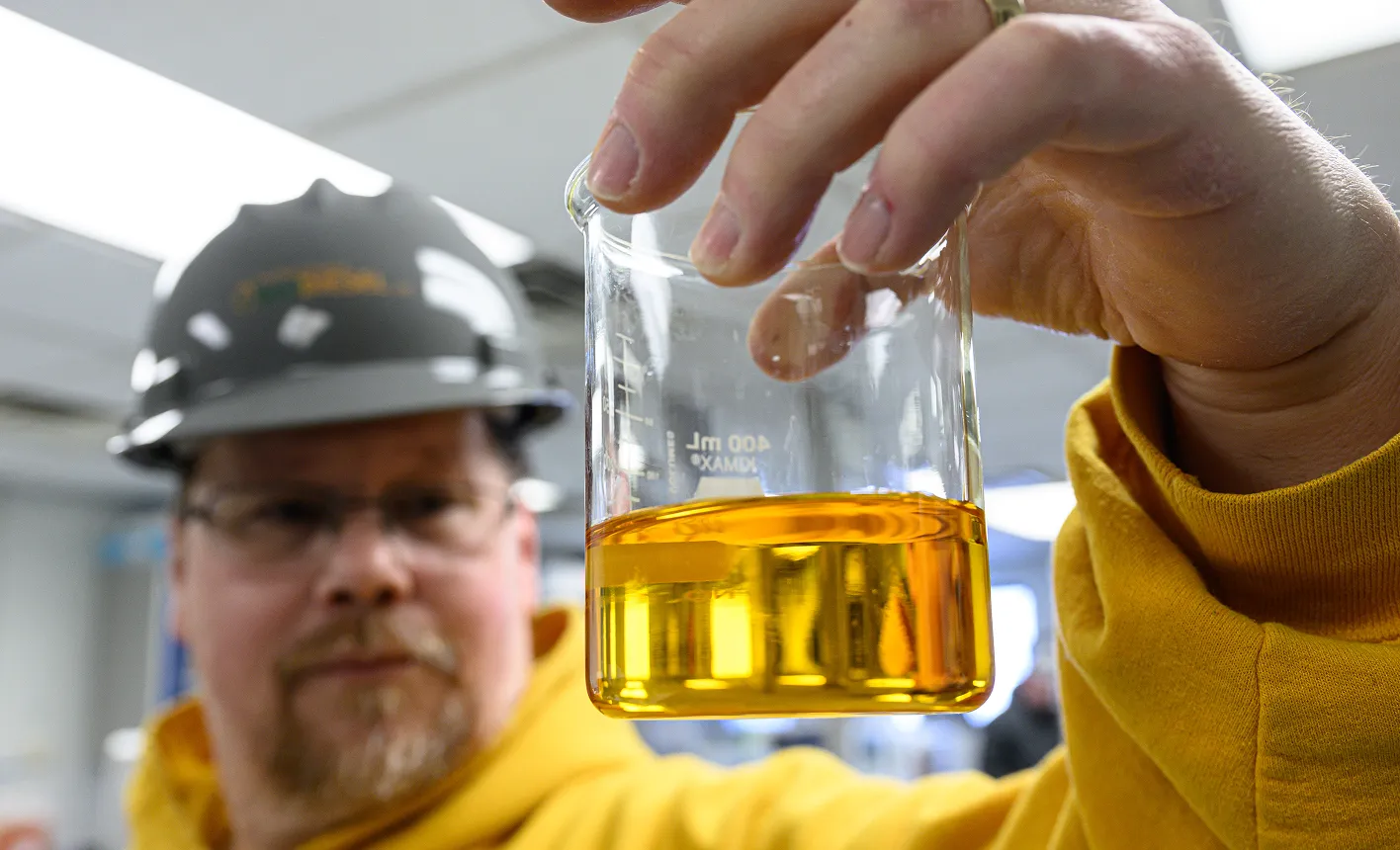 Person in protective workwear holding a beaker of soybean oil for inspection, with an industrial or laboratory setting in the background.